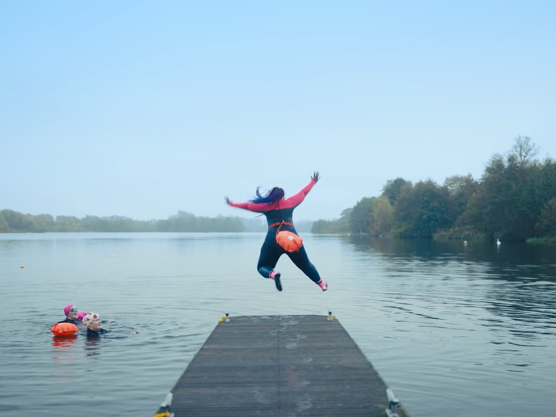 Person in a wetsuit leaps off a wooden dock into a calm lake while others in life jackets swim nearby.