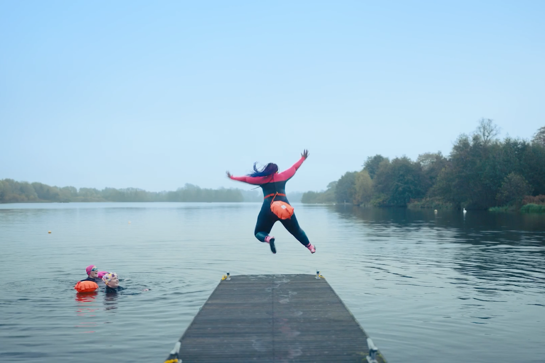 Person in a wetsuit leaps off a wooden dock into a calm lake while others in life jackets swim nearby.