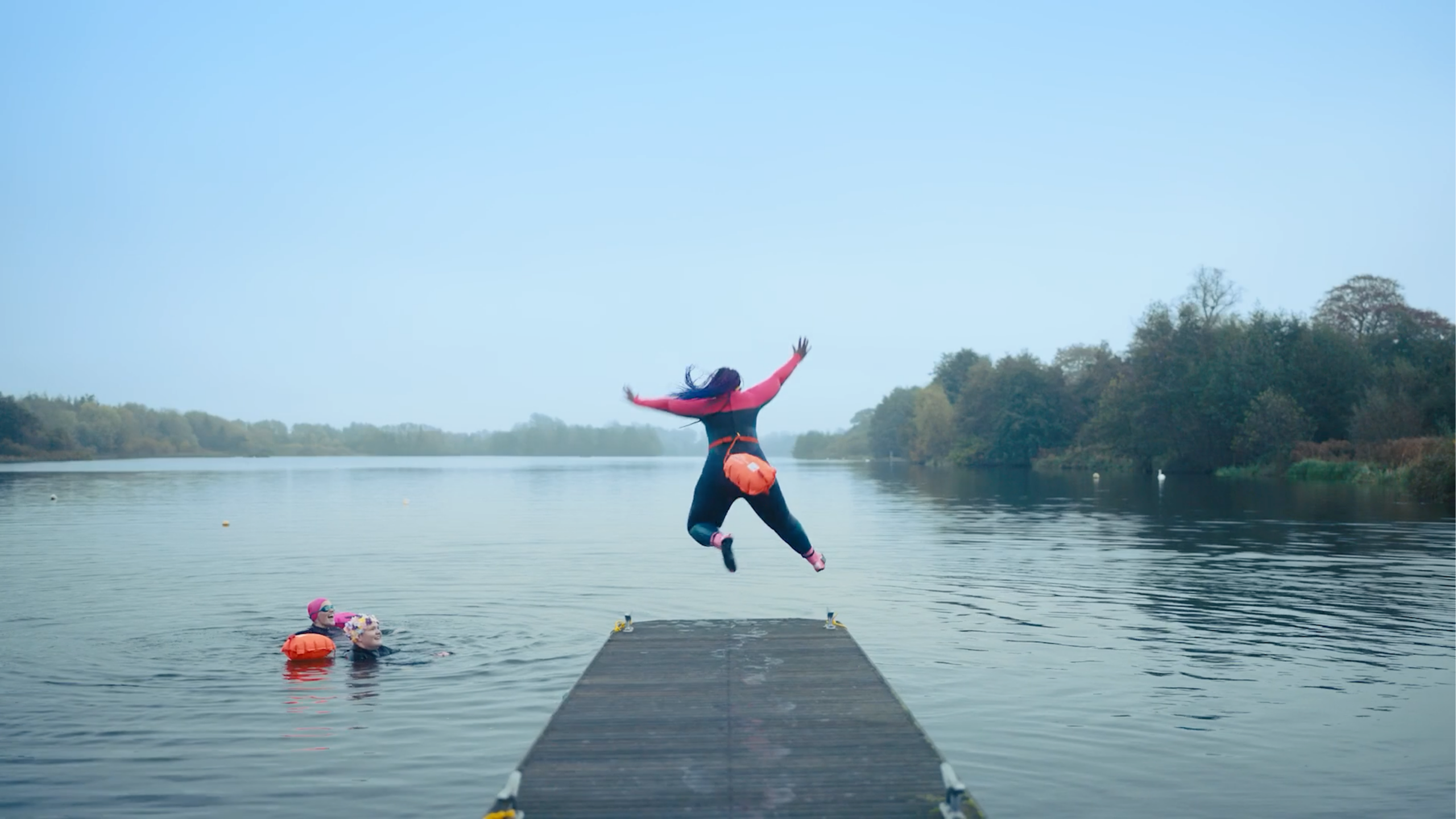 Person in a wetsuit leaps off a wooden dock into a calm lake while others in life jackets swim nearby.