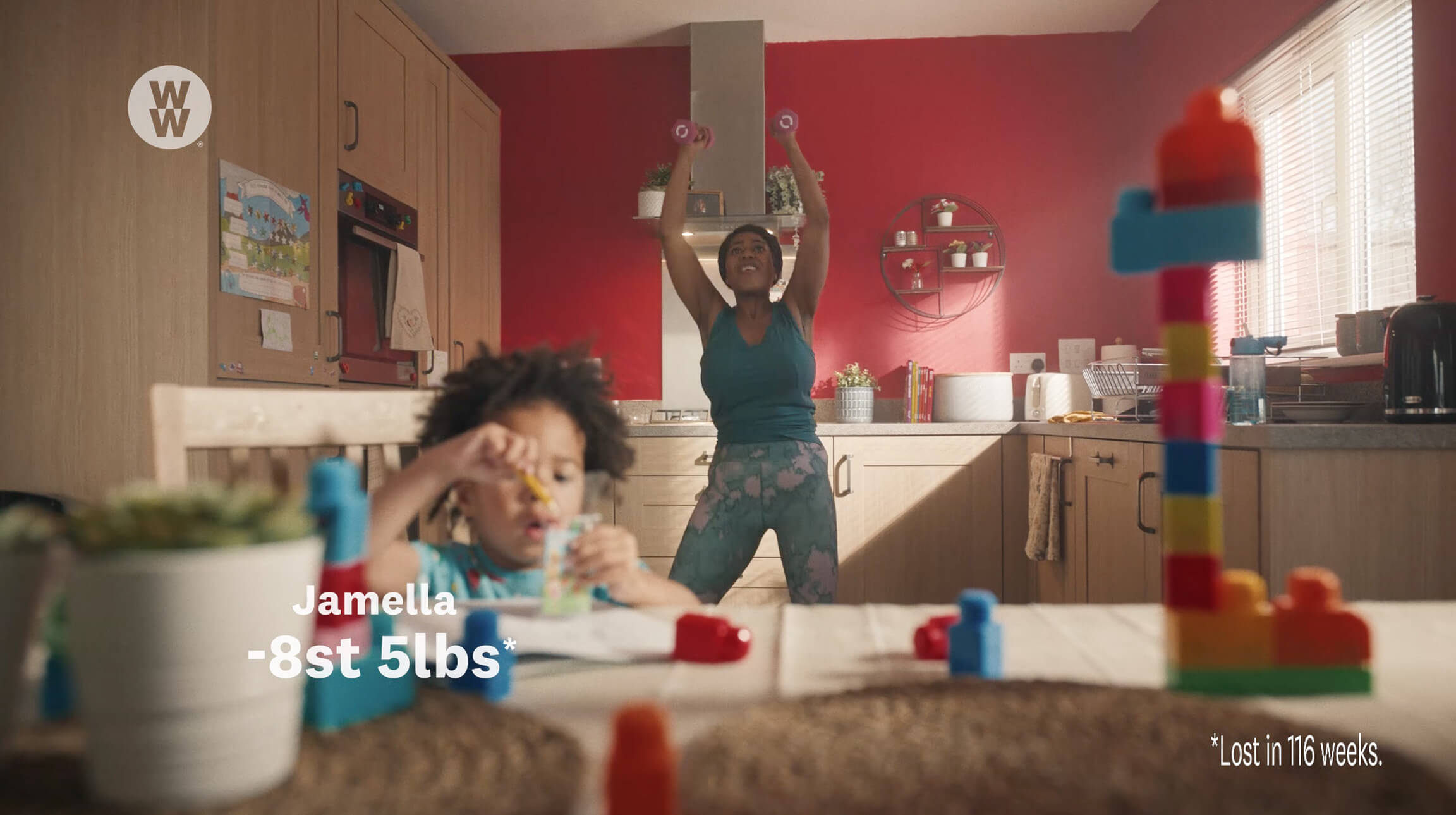 Woman in teal workout clothes lifts dumbbells overhead in a red kitchen as a child plays with blocks in the foreground; WW logo on the cabinet visible.